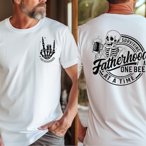 Puede incluir: Camiseta blanca con un gr&aacute;fico de mano de esqueleto negro haciendo un gesto de rock and roll, y la palabra "FATHERHOOD". La parte posterior de la camiseta presenta un esqueleto bebiendo cerveza con el texto "Surviving Fatherhood One Beer At A Time."