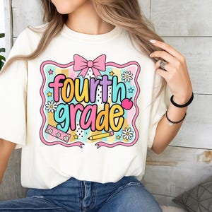 Op de afbeelding: Cr&egrave;mekleurig T-shirt met een kleurrijke afbeelding met de tekst "fourth grade". Het ontwerp bevat een roze strik, madeliefjes, sterren, een liniaal, een potlood en een appel. De tekst is in een mix van kleuren, waaronder geel, blauw, roze en paars.