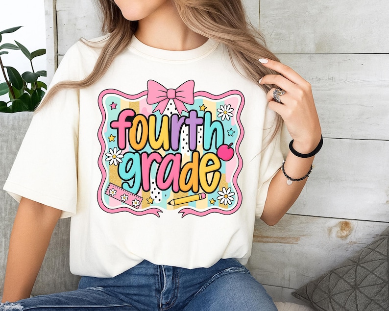 Op de afbeelding: Cr&egrave;mekleurig T-shirt met een kleurrijke afbeelding met de tekst "fourth grade". Het ontwerp bevat een roze strik, madeliefjes, sterren, een liniaal, een potlood en een appel. De tekst is in een mix van kleuren, waaronder geel, blauw, roze en paars.