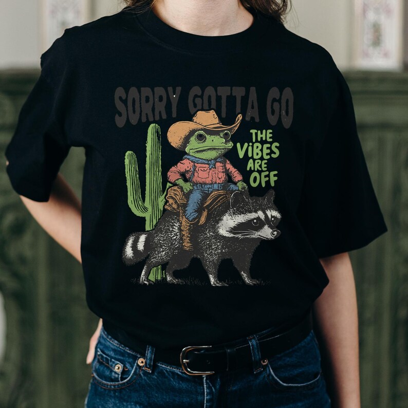 Może przedstawiać: Czarny t-shirt z grafiką przedstawiającą zieloną żabę w brązowym kowbojskim kapeluszu i czerwonej koszuli, jadącą na szopie praczu. Obok żaby znajduje się zielony kaktus. Napis na koszulce brzmi SORRY GOTTA GO oraz THE VIBES ARE OFF.