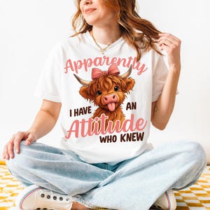 May include: White t-shirt featuring a cartoon Highland cow with a pink bow and the text "Apparently I have an attitude who knew" in pink script. The cow has brown fur and is sticking out its tongue.