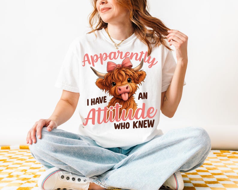 May include: White t-shirt featuring a cartoon Highland cow with a pink bow and the text "Apparently I have an attitude who knew" in pink script. The cow has brown fur and is sticking out its tongue.