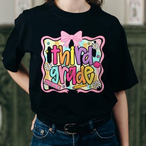 Puede incluir: Una camiseta negra con un gr&aacute;fico colorido que incluye el texto "third grade" en letras de burbuja. El dise&ntilde;o presenta un lazo rosa, una regla, un l&aacute;piz, una manzana y margaritas blancas sobre un fondo de acuarela pastel con estrellas.