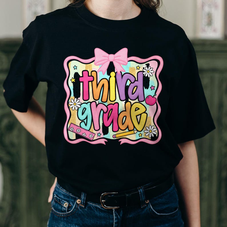 Puede incluir: Una camiseta negra con un gr&aacute;fico colorido que incluye el texto "third grade" en letras de burbuja. El dise&ntilde;o presenta un lazo rosa, una regla, un l&aacute;piz, una manzana y margaritas blancas sobre un fondo de acuarela pastel con estrellas.
