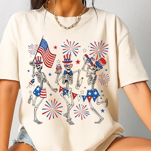 May include: Cream-colored t-shirt featuring a festive skeleton design. Three skeletons are depicted celebrating with an American flag, fireworks, and patriotic accessories. The design incorporates red, white, and blue colors, perfect for Independence Day.