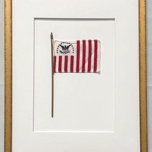 May include: A framed historical flag featuring red and white vertical stripes with a blue canton containing a black eagle emblem surrounded by a semi-circle of white stars. The flag is mounted on a thin pole and enclosed in a gold-toned frame.