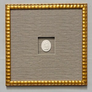 May include: A square wall art piece featuring a central white oval cameo with a relief design, mounted on a textured light gray fabric background. The artwork is enclosed in a decorative gold frame with a scalloped, repeating edge pattern.