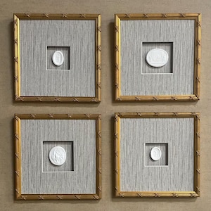 May include: Four framed wall art pieces. Each features a gold-colored bamboo-style frame, surrounding a square of gray fabric. Centered within each frame is a white, oval-shaped relief sculpture. The sculptures depict classical scenes and figures.