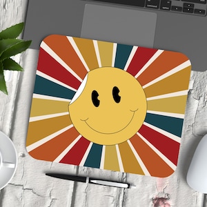Puede incluir: Una cara sonriente amarilla con un diseño retro de rayos de sol en colores rojo, naranja, amarillo y azul. La cara sonriente es una pegatina con una esquina despegada. El fondo es una superficie blanca.