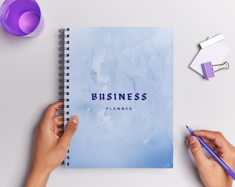 May include: A spiral-bound business planner with a light blue watercolor design. The cover features the word "BUSINESS" above "PLANNER" in a dark blue font. A purple pen and a purple cup are also visible.