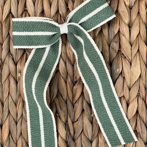 May include: A green and white striped bow made of woven fabric. The bow has a white center knot and long, flowing tails. The bow is set against a textured, woven background.