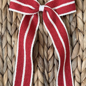 May include: A decorative bow featuring a red and white striped pattern. The bow is made of a textured fabric and has long, flowing tails. The bow is tied with a white ribbon in the center. The background is a woven texture.