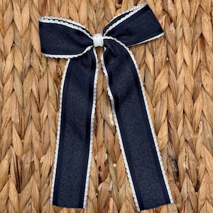 May include: A navy blue bow with white scalloped edges and a white center knot. The bow has long, flowing tails and is set against a woven background. This decorative bow is suitable for hair accessories or gift wrapping.