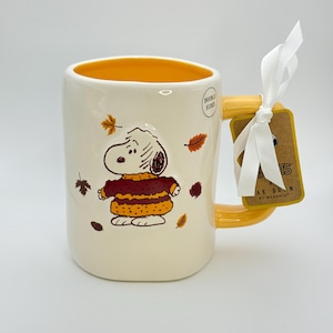 May include: Cream-colored ceramic mug featuring Snoopy in a sweater, with autumn leaves. The mug has a yellow handle and interior, and a tag tied with a white ribbon. The text on the tag reads "Rae Dunn by Magenta."