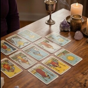 May include: A wooden table displays a tarot card spread with various illustrations. A silver chalice, lit candle, and amethyst crystals are also present. Wisps of smoke rise from the chalice, creating a mystical atmosphere.