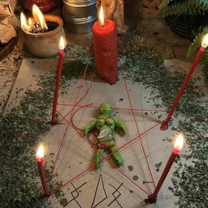 May include: A ritualistic scene featuring a green figurine at the center, encircled by red string and four lit red candles. A large red candle stands nearby. The scene is set on a surface covered with dried herbs, with additional candles and a statue in the background.