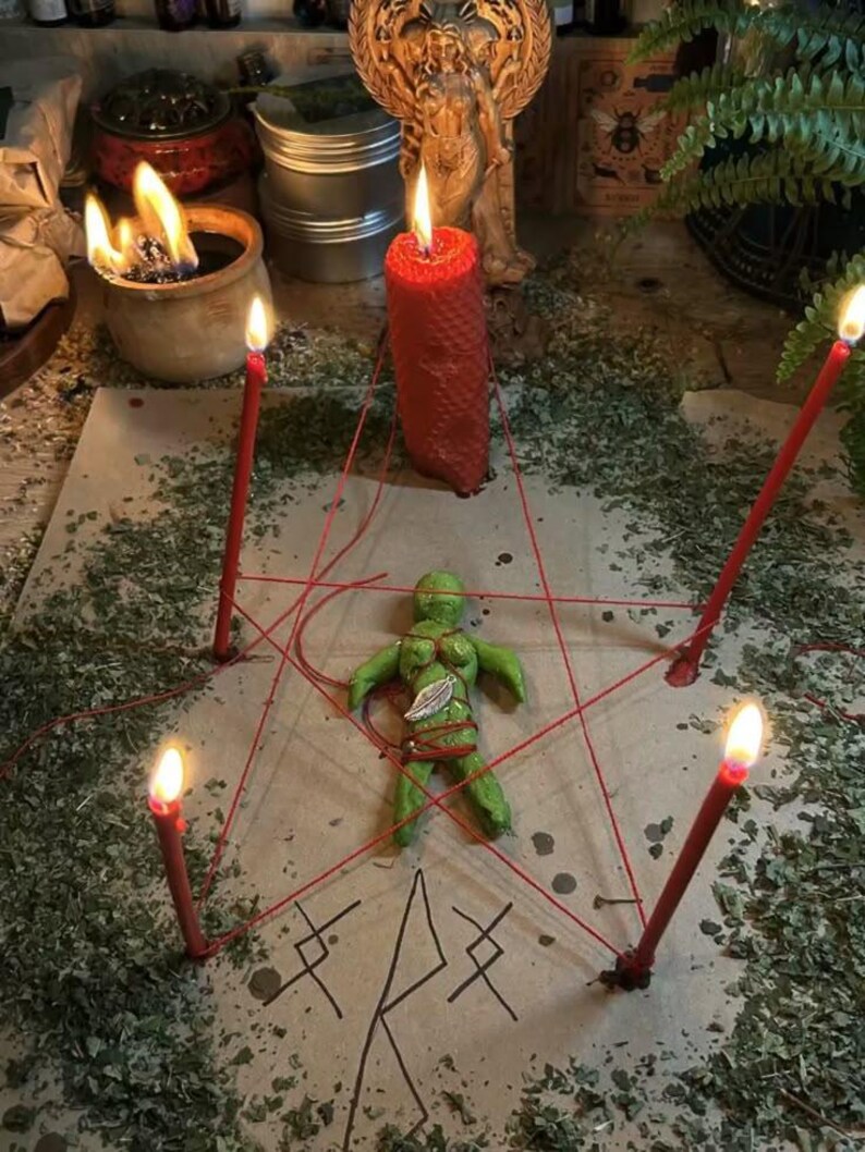 May include: A ritualistic scene featuring a green figurine at the center, encircled by red string and four lit red candles. A large red candle stands nearby. The scene is set on a surface covered with dried herbs, with additional candles and a statue in the background.