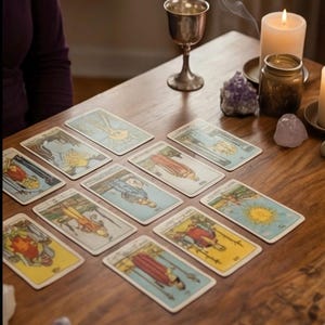 May include: A wooden table displays a tarot card spread with various illustrations. A silver chalice, lit candle, and amethyst crystals are also present. Wisps of smoke rise from the chalice, creating a mystical atmosphere.