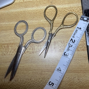 May include: Two pairs of decorative scissors with ornate handles and pointed blades. One pair is silver-toned, the other is gold-toned. A white measuring tape with black markings and the text "60 inch" is also present.