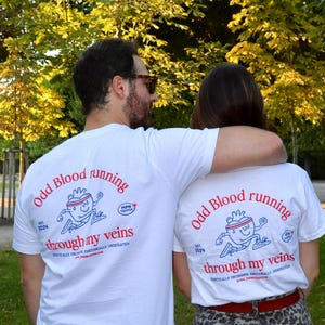 Peut inclure: Deux personnes portant des t-shirts blancs avec le texte "Odd Blood running through my veins" en rouge. Les chemises présentent un personnage de cœur de dessin animé et le texte "MPN Fighter". Les chemises sont blanches.
