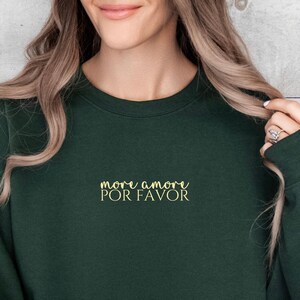 Pode incluir: Um moletom verde escuro com a frase "more amore por favor" em letras douradas. O moletom é de gola redonda e tem mangas compridas. O texto está centralizado no peito.