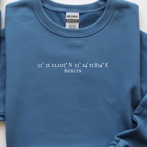 May include: A blue crew neck sweatshirt with the text "52° 31' 12.025" N 13° 24' 17.834" E BERLIN" printed in white. The sweatshirt features a classic design with a ribbed neckline, cuffs, and hem. The Gildan brand label is visible.