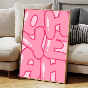 May include: A framed art print with the word "YEAH" in large, bubble-style pink letters. The letters have a glossy, highlighted effect, and the background is a lighter shade of pink. The frame is a light wood color.