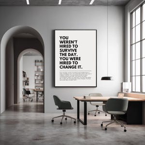 Puede incluir: Un p&oacute;ster motivacional enmarcado con el texto "YOU WEREN'T HIRED TO SURVIVE THE DAY. YOU WERE HIRED TO CHANGE IT." se muestra en un entorno de oficina moderno. El p&oacute;ster es en blanco y negro y cuelga sobre un escritorio de madera.