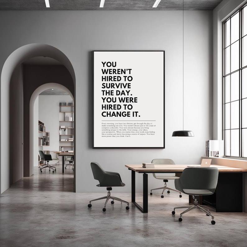 Puede incluir: Un p&oacute;ster motivacional enmarcado con el texto "YOU WEREN'T HIRED TO SURVIVE THE DAY. YOU WERE HIRED TO CHANGE IT." se muestra en un entorno de oficina moderno. El p&oacute;ster es en blanco y negro y cuelga sobre un escritorio de madera.