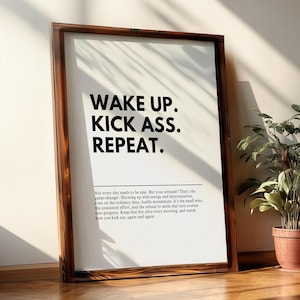 May include: A framed print with the text "WAKE UP. KICK ASS. REPEAT." in bold black letters. The print is in a dark brown wooden frame. Below the text is a paragraph of text in a smaller font. A potted plant is in the background.
