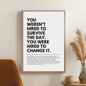 Puede incluir: Un cartel enmarcado con el texto "YOU WEREN'T HIRED TO SURVIVE THE DAY. YOU WERE HIRED TO CHANGE IT." en letras negras en negrita sobre un fondo blanco. El cartel est&aacute; en un marco marr&oacute;n claro. El cartel tambi&eacute;n incluye un p&aacute;rrafo de texto.
