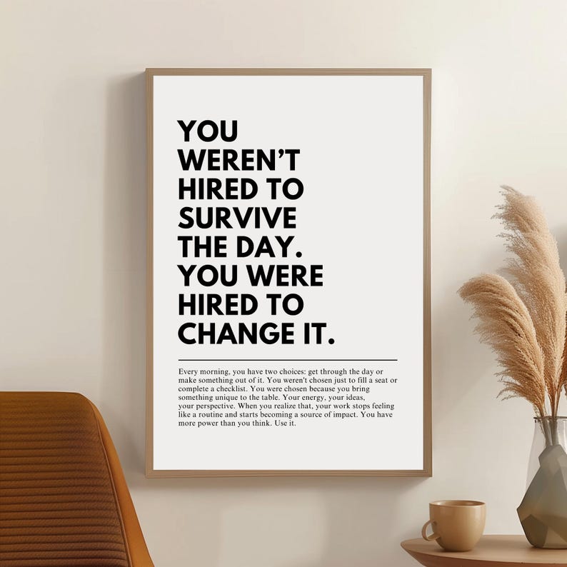 Puede incluir: Un cartel enmarcado con el texto "YOU WEREN'T HIRED TO SURVIVE THE DAY. YOU WERE HIRED TO CHANGE IT." en letras negras en negrita sobre un fondo blanco. El cartel est&aacute; en un marco marr&oacute;n claro. El cartel tambi&eacute;n incluye un p&aacute;rrafo de texto.