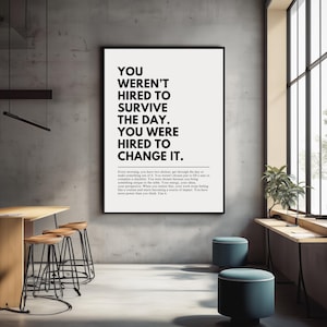 Puede incluir: Un p&oacute;ster motivacional en blanco y negro enmarcado con el texto "YOU WEREN'T HIRED TO SURVIVE THE DAY. YOU WERE HIRED TO CHANGE IT." El p&oacute;ster se muestra en un entorno de oficina moderno con taburetes y una mesa.
