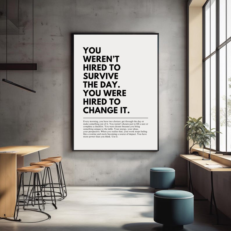 Puede incluir: Un p&oacute;ster motivacional en blanco y negro enmarcado con el texto "YOU WEREN'T HIRED TO SURVIVE THE DAY. YOU WERE HIRED TO CHANGE IT." El p&oacute;ster se muestra en un entorno de oficina moderno con taburetes y una mesa.