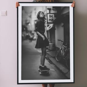 Può includere: Una fotografia in bianco e nero di una donna che fa skateboard in una strada. Indossa una gonna nera e una camicia nera. La foto è scattata da un basso angolo, e la donna guarda la fotocamera. C'è un cartello che dice "B4 PUB" sullo sfondo.