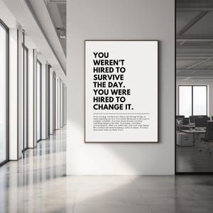 Puede incluir: Un p&oacute;ster enmarcado con el texto "YOU WEREN'T HIRED TO SURVIVE THE DAY. YOU WERE HIRED TO CHANGE IT." en un entorno de oficina moderno. El p&oacute;ster tiene un fondo blanco con texto negro.