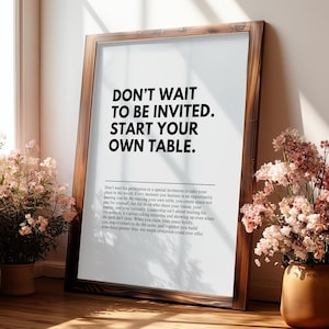 May include: A framed print with the text "DON'T WAIT TO BE INVITED. START YOUR OWN TABLE." The wooden frame surrounds a white background with additional text below the main message. Pink flowers in a vase are in the background.