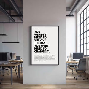 Puede incluir: P&oacute;ster enmarcado en negro con el texto "YOU WEREN'T HIRED TO SURVIVE THE DAY. YOU WERE HIRED TO CHANGE IT." en una oficina moderna. El p&oacute;ster es en blanco y negro y est&aacute; colgado en una pared blanca.