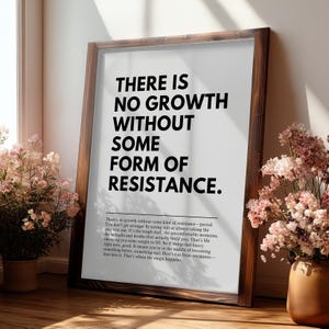 May include: A framed print with the text "THERE IS NO GROWTH WITHOUT SOME FORM OF RESISTANCE." The print is in a dark brown wooden frame. The text is in a bold, sans-serif font. The print is leaning against a wall near a window.