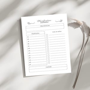May include: A white daily planner with the text "Planificateur Journalier" and sections for daily planning, to-do lists, and gratitude. The planner includes time slots from 5 am to 5 pm. The design is simple and elegant, with a minimalist aesthetic.
