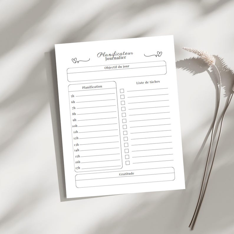 May include: A white daily planner with the text "Planificateur Journalier" and sections for daily planning, to-do lists, and gratitude. The planner includes time slots from 5 am to 5 pm. The design is simple and elegant, with a minimalist aesthetic.