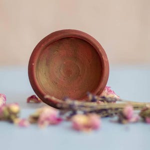 May include: A small, round, terracotta-colored bowl with a textured interior. The bowl is surrounded by dried lavender and pink rose buds, creating a rustic and natural aesthetic. The bowl is likely used for decorative purposes.