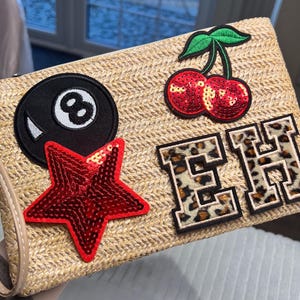 May include: A beige woven clutch bag with a tan handle. The bag is decorated with a black eight ball patch, a red sequined star, a pair of red sequined cherries, and two leopard print letter patches spelling "EH".