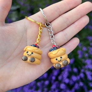 May include: Two keyrings with miniature pancake stacks, each with a smiling face, blueberries, and butter. One has a gold chain, the other a silver chain. The pancake stacks have brown feet and are held by a hand.