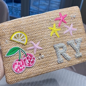 May include: A rectangular straw clutch bag featuring embroidered patches of a lemon slice, cherries, and starfish. The letters "RY" are decorated with pearls. Scattered pearls add a touch of elegance.