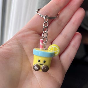 May include: A novelty keychain featuring a cartoon lemonade drink design. The keychain is yellow with a blue rim, brown 'feet', and a smiling face. It includes a pink and white striped straw and a lemon slice. Attached to a silver metal ring.