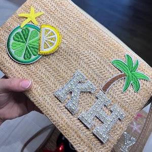 May include: A woven straw clutch bag with a zip. It features embroidered patches of a lime, lemon, starfish, and palm tree. The initials "KH" are spelled out with pearl-like beads.