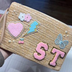 May include: A straw-colored clutch with a zipper closure and a wrist strap. The clutch is decorated with embroidered patches including a pink heart, a blue lightning bolt, and the letters "ST". Additional embellishments include pearls and floral and butterfly patches.