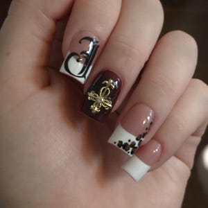 Puede incluir: Primer plano de una mano con uñas manicuradas. Una uña presenta un diseño en blanco y negro con la letra "J". Otra uña es de color burdeos con un adorno dorado. Las uñas restantes tienen una punta blanca con motas negras.