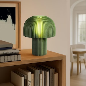 May include: A green mushroom-shaped table lamp with a ribbed shade and cylindrical base. The lamp is made of a textured material and sits on a wooden shelf. The lamp is illuminated, casting a warm glow.